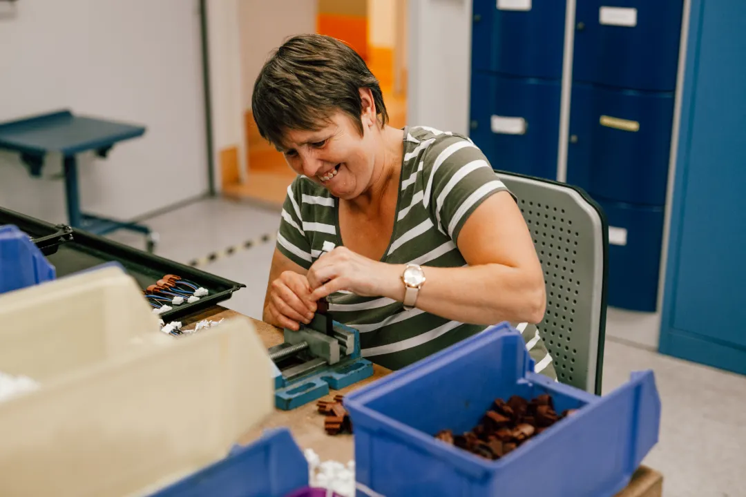 Eine lächelnde Frau sitzt an einem Werktisch und montiert kleine Bauteile in einer Werkstattumgebung.