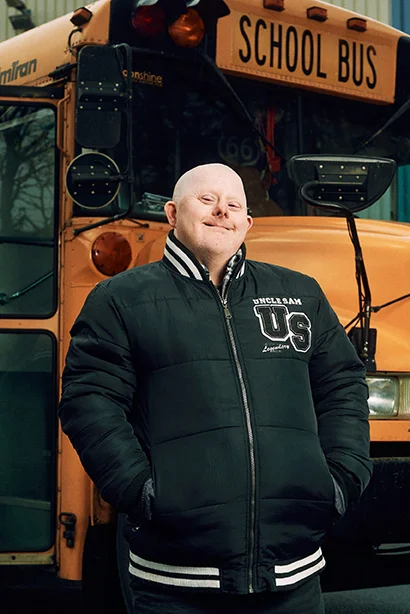 Person steht selbstbewusst vor einem gelben Schulbus und trägt eine schwarze Jacke.