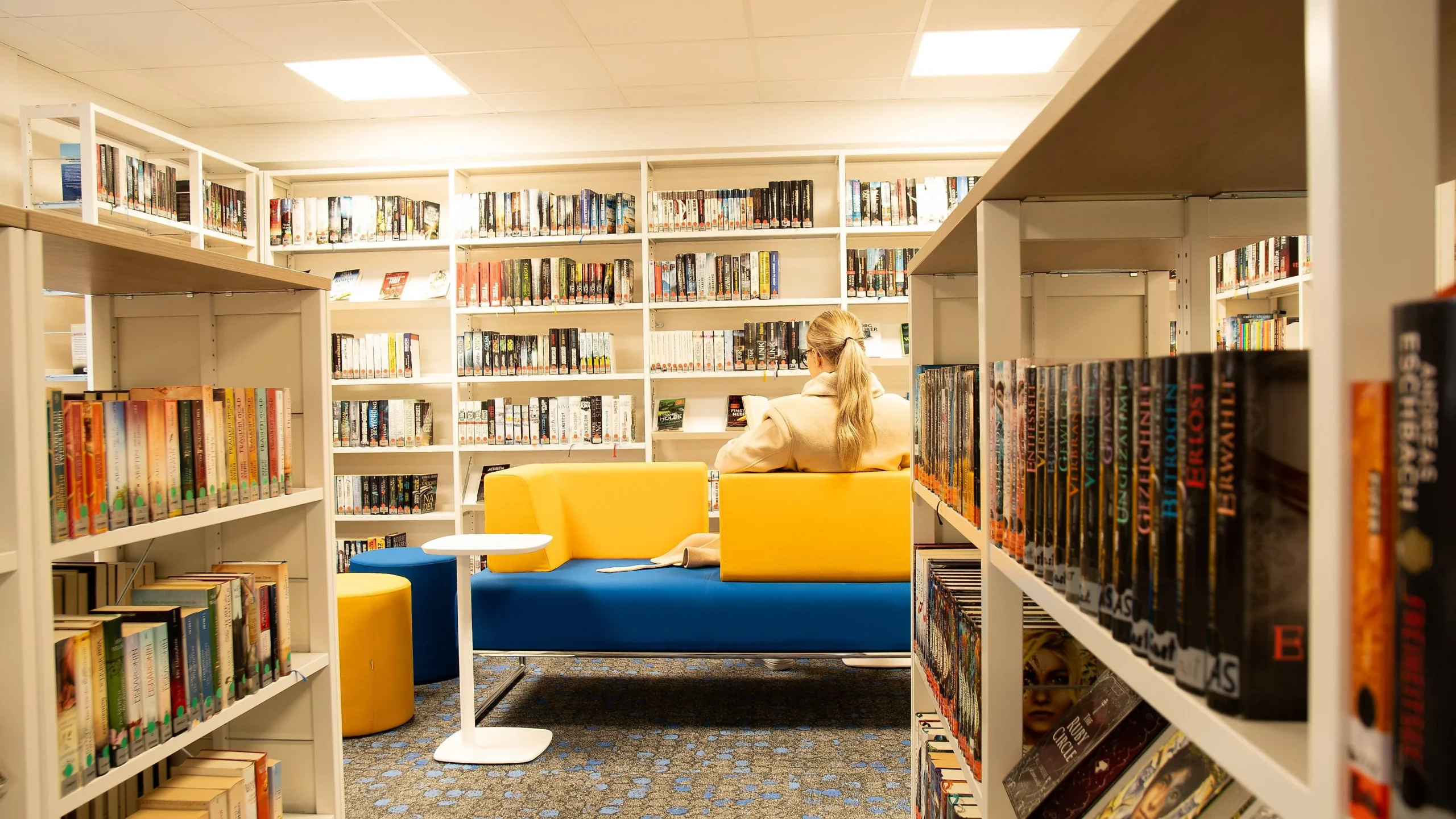 „Innenraum einer modernen Bibliothek mit hell beleuchteten Regalen voller Bücher. In der Mitte sitzt eine Person mit blondem, zu einem Pferdeschwanz gebundenem Haar und beigefarbener Jacke auf einem gelb-blauen Sofa und liest. Links und rechts stehen hohe Bücherregale; der Raum wirkt ruhig und einladend.