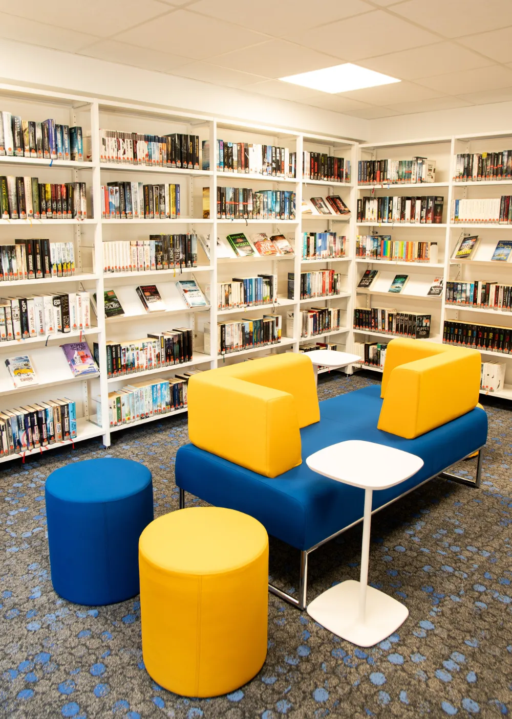 Sitzbereich mit gelb-blauem Sofa und runden Hockern vor vollen Bücherregalen.