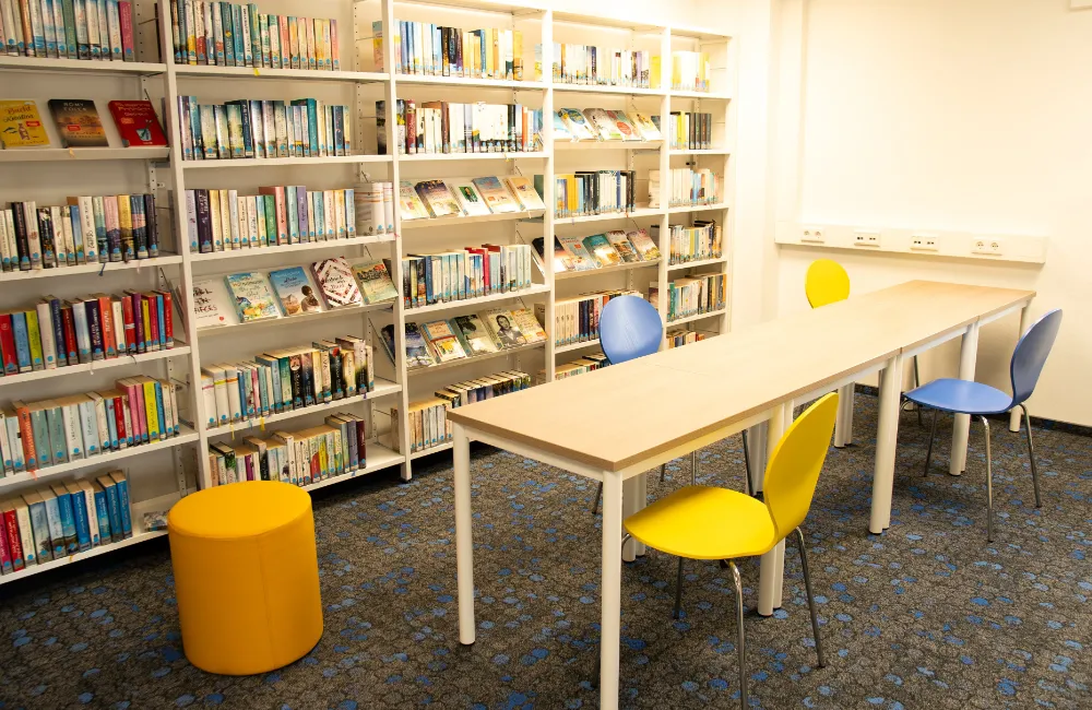 Langer Tisch mit bunten Stühlen vor Regalen voller Bücher.