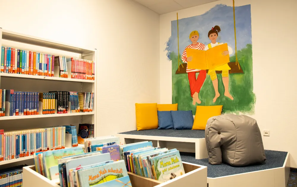 Kinderbereich der Bibliothek mit Bücherregalen, Sitzstufen und Wandbild zweier Kinder.
