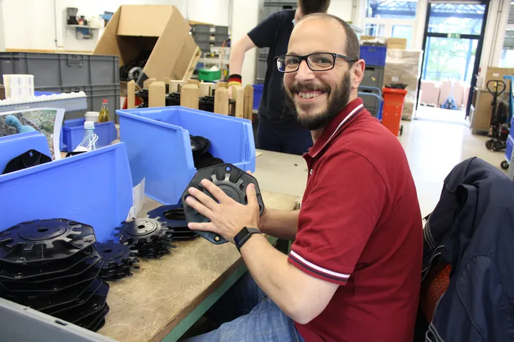 Ein Mann sitzt in einer Werkstatt und hält ein Werkzeug in den Händen. Er trägt ein schwarzes Shirt mit dem Aufdruck „Never too old to rock“ und lächelt. Um ihn herum liegen Werkmaterialien und Sortierboxen.